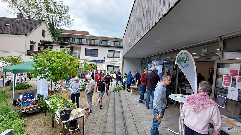 Menschen auf einem Gehweg vor einem Gebäude mit Informationsständen und einem Banner mit grünem Logo