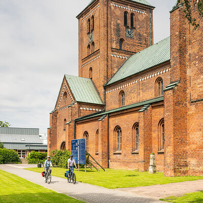 Bild vergrößern: Zwei Radfahrende fahren auf einem Weg vor einer großen Backstein-Kirche.